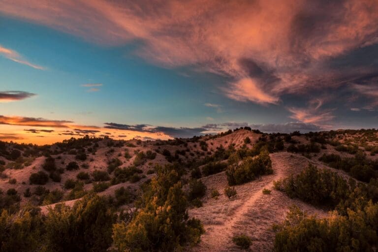 A vibrant sunset over desert hills, with dramatic orange and pink clouds contrasting a deep blue sky and scattered greenery.