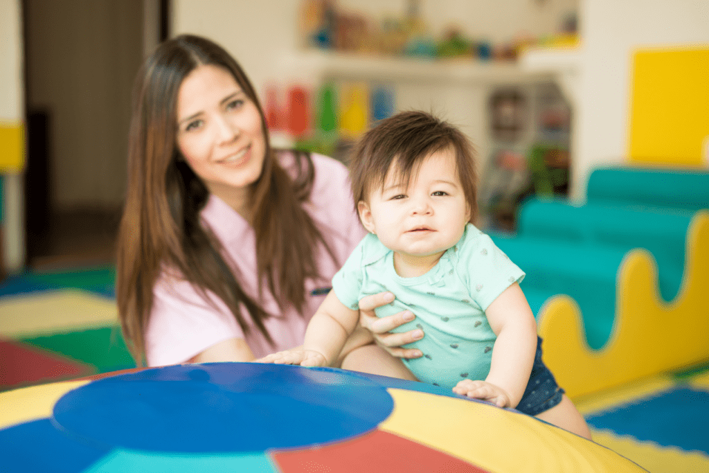 A woman holds a baby leaning on a colorful play mat in a bright indoor play area.