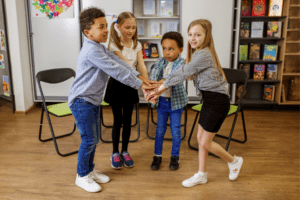 Four children standing together in a classroom, forming a circle with their hands stacked