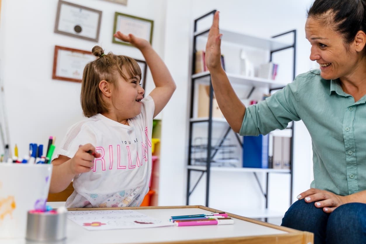 Smiling ABA therapist celebrates a learning milestone with a young girl during a therapy activity in a well-equipped center.