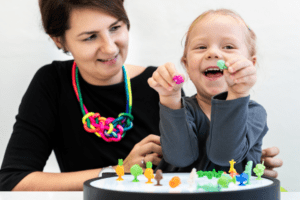 A smiling child holds colorful toy figurines while sitting next to a supportive adult during a playful learning activity.
