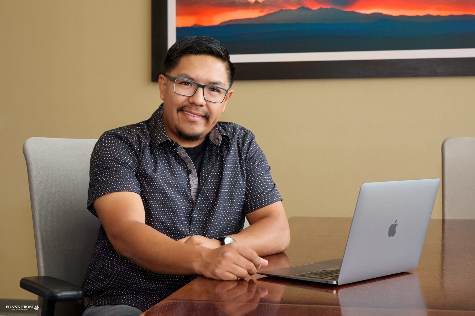 A man with glasses and short black hair wearing a dark blue shirt sits at a wooden desk with a laptop open. He faces the camera and smiles.