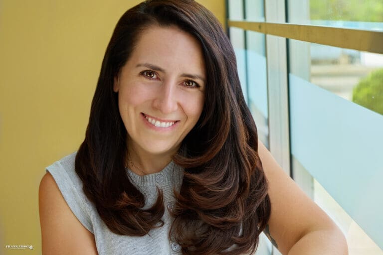 A woman with long dark brown hair and a light blue shirt smiles next to a brightly lit window with an obscured view of a tree.
