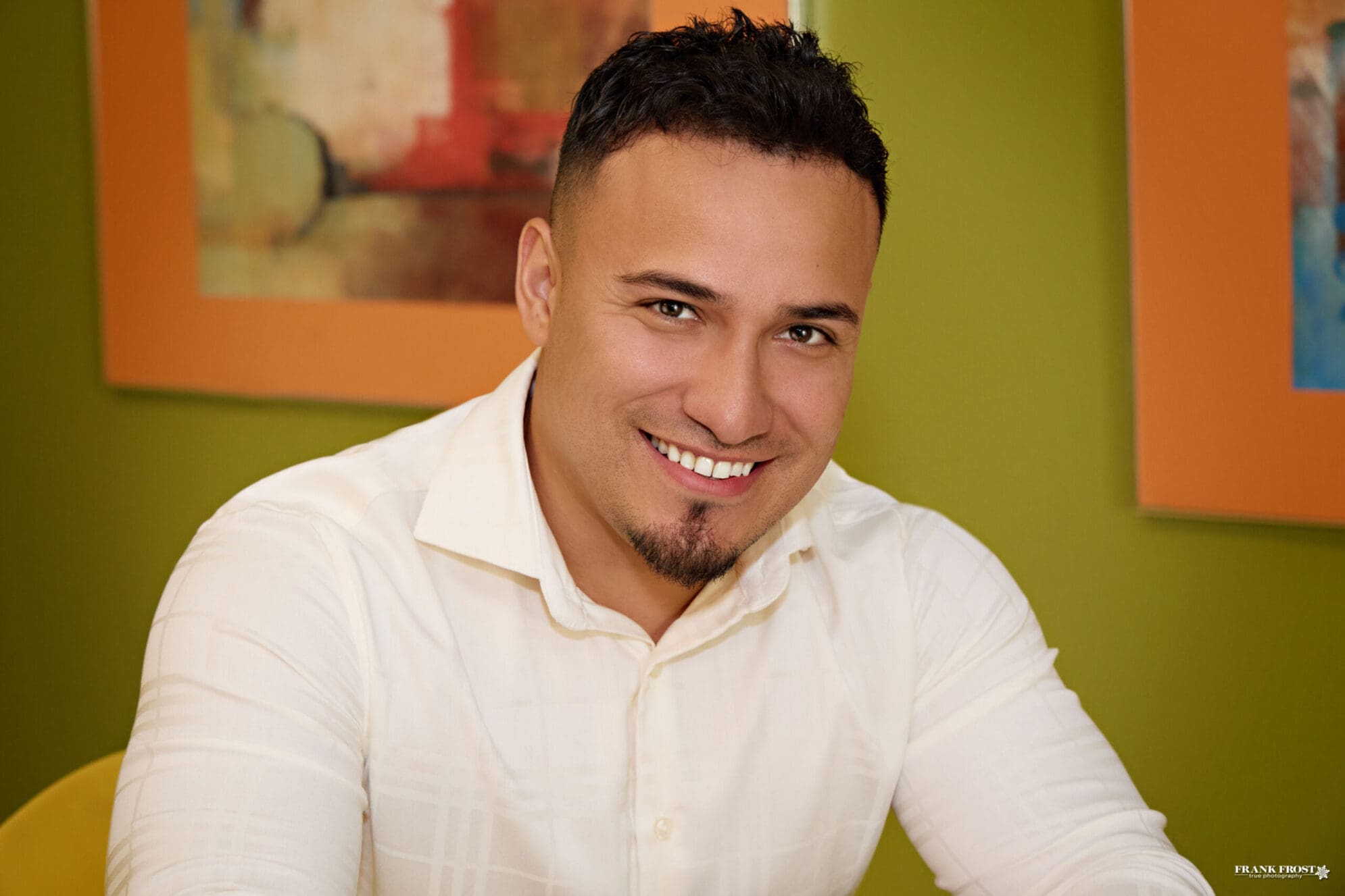A man with short black hair, a goatee and a white button down shirt smiles in front of an olive green wall with blurred photos in frames.