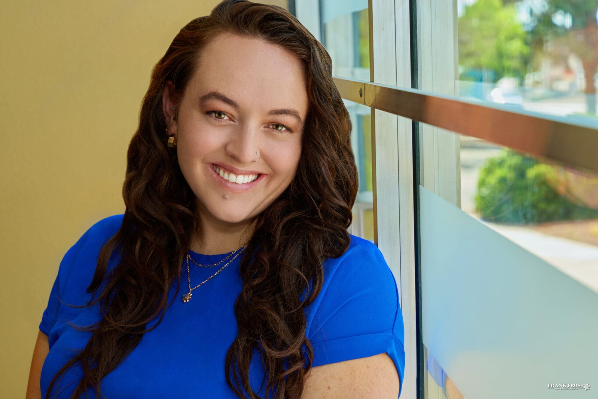 A woman with long brown hair parted to the side wearing a bright blue shirt smiles next to a large tinted window with a view of a tree.