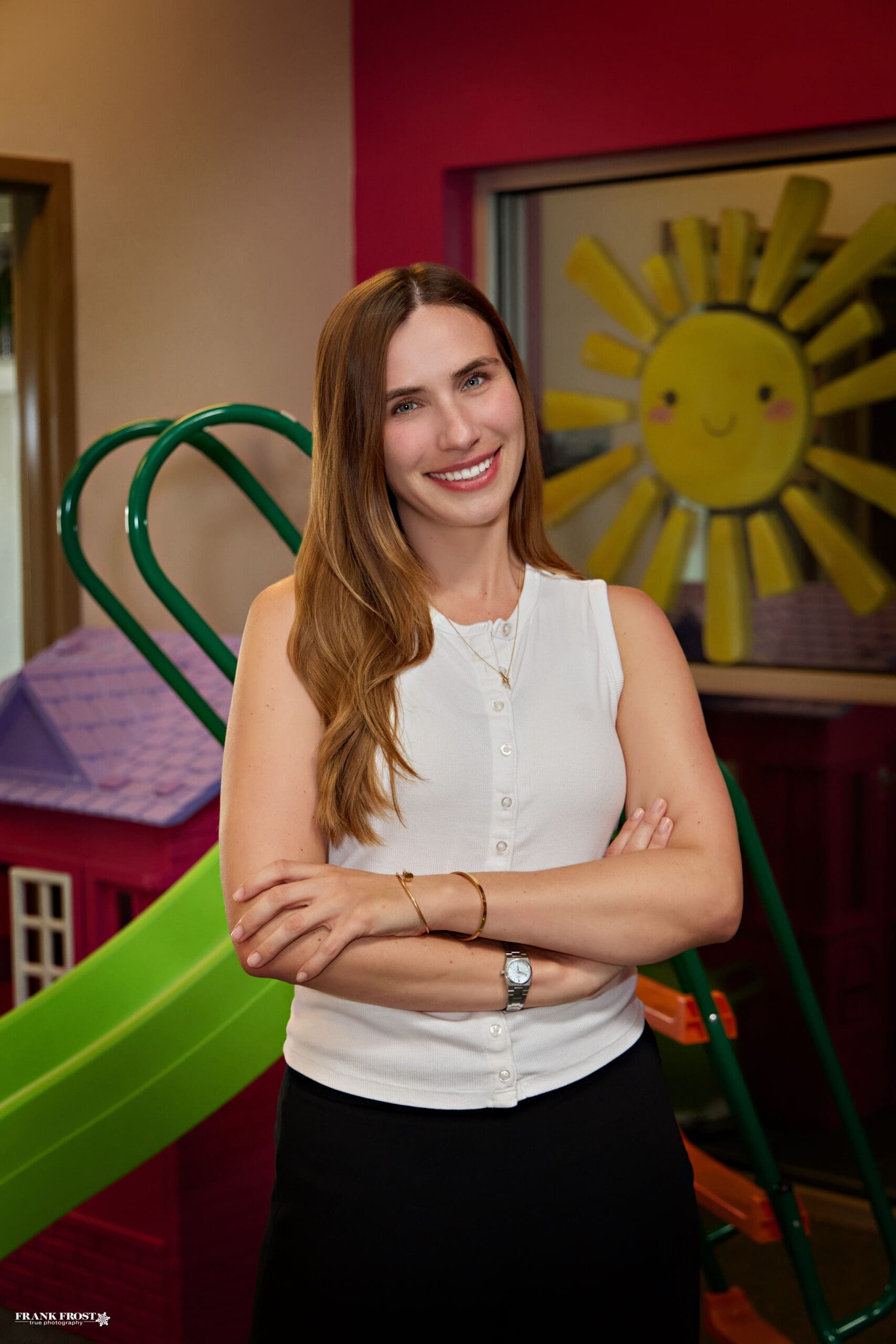 A woman in a white tank top and long light brown hair smiles brightly with blurred indoor playground equipment behind her.