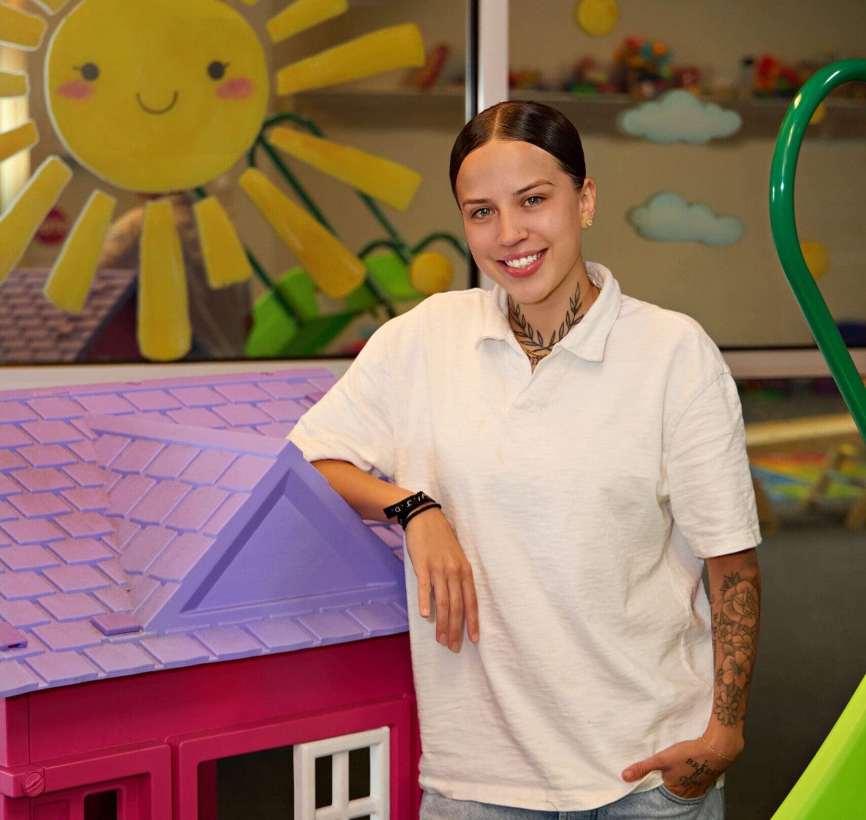 A woman with her hair in a low ponytail and white oversized polo shirt rests one arm on a large pink dollhouse, the other arm, sporting a sleeve tattoo, rests with her hand in her front pants pocket. She smiles brightly.