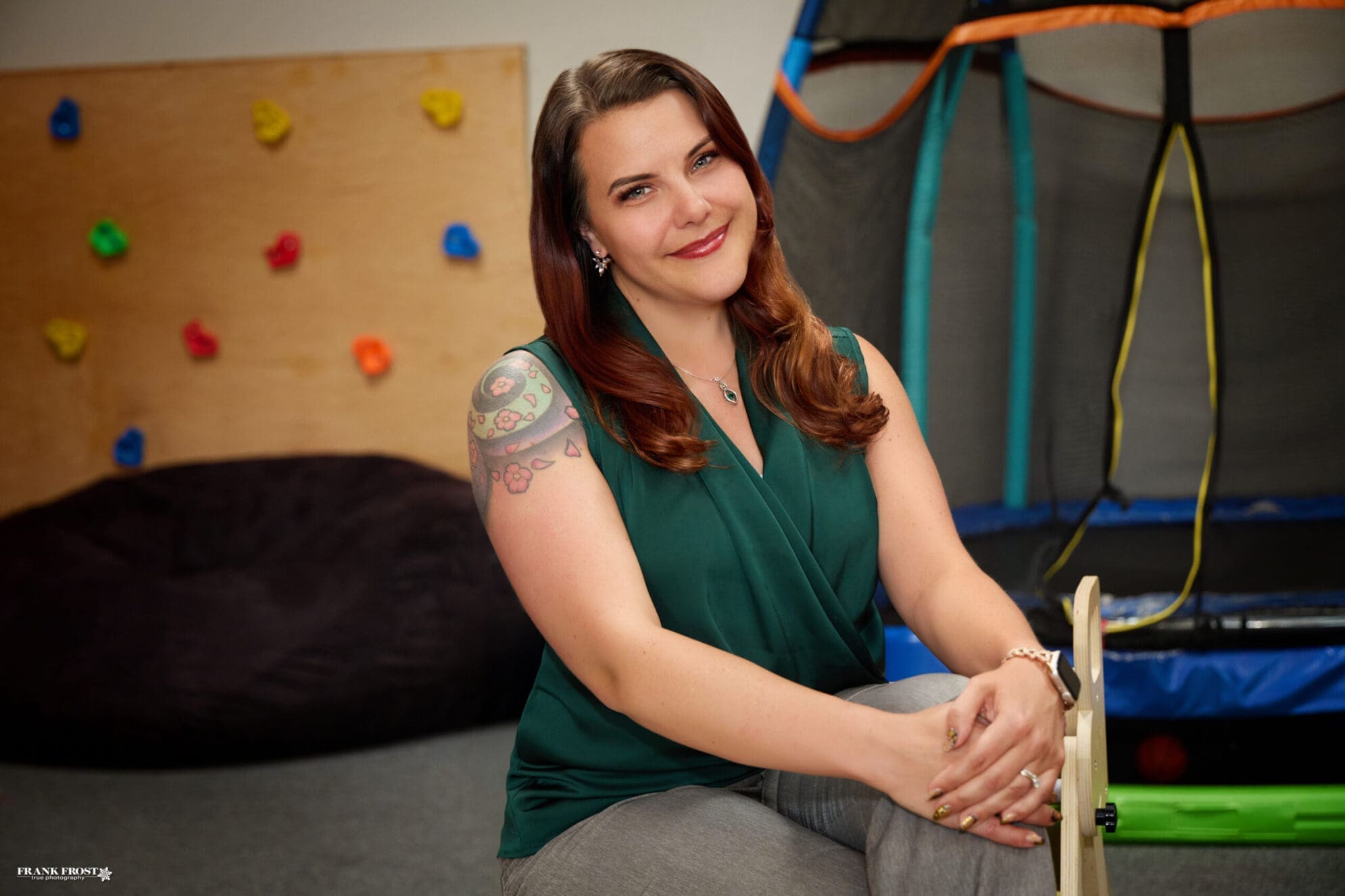 A woman with auburn hair parted on the side in a dark green flowy top and a large shoulder tattoo sits crossed legged with her hands folded over her left knee. The background shows a mini rock wall and an indoor trampoline.