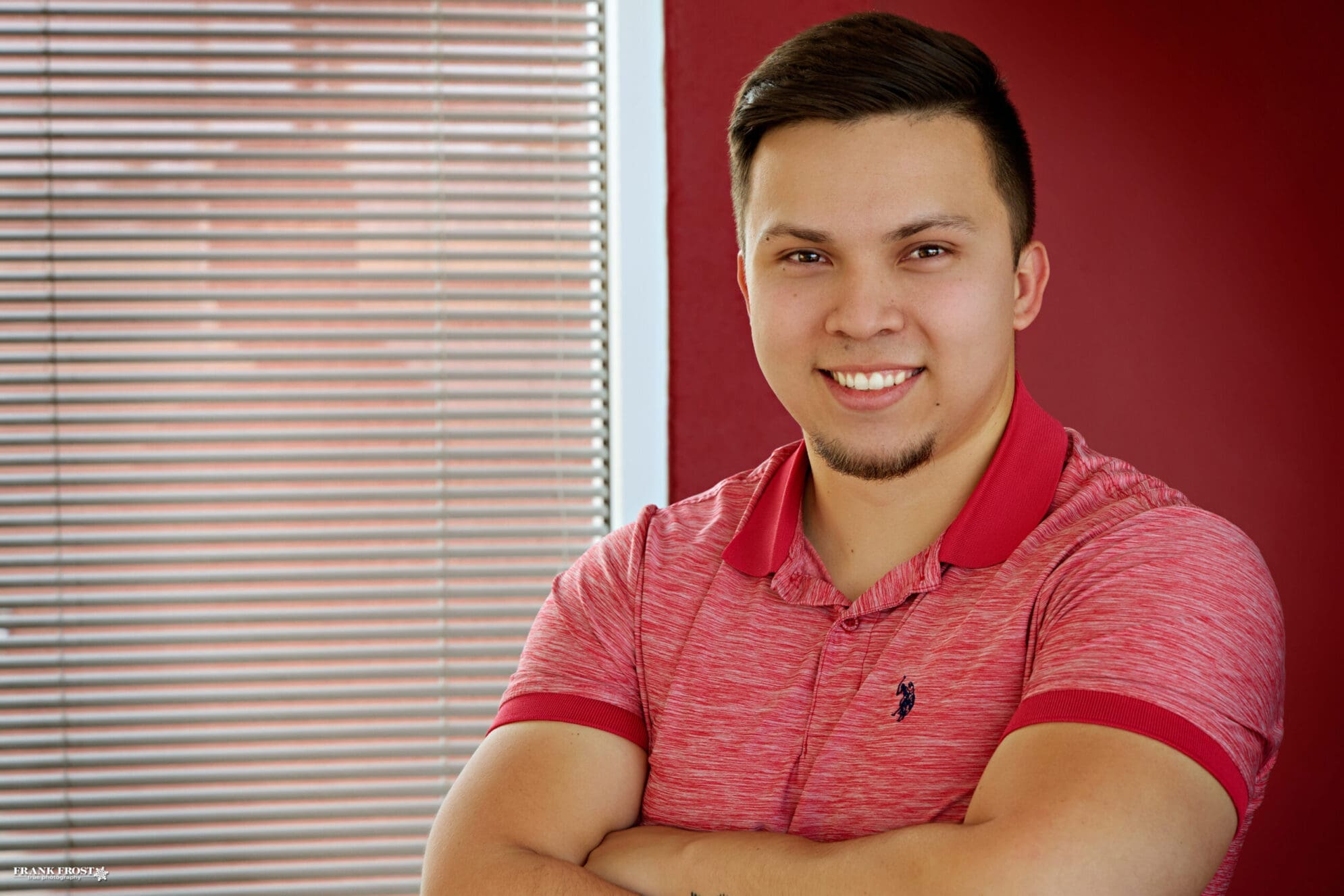 A man with short black hair and a red polo shirt stands smiling with his arms crossed confidently in front of a red wall and a window with the blinds closed.